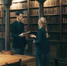 a man and woman standing in front of a library full of books, talking to each other