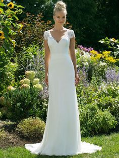 a woman in a white wedding dress standing on the grass with flowers behind her and looking at the camera