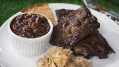 a white plate topped with ribs and beans next to bread on top of a green grass covered field