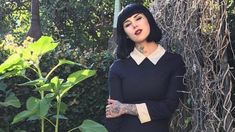 a woman with black hair and tattoos standing in front of a bush wearing a black dress