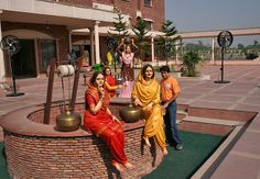 three women in sari sitting on a brick fountain with bells around it and two men standing next to them