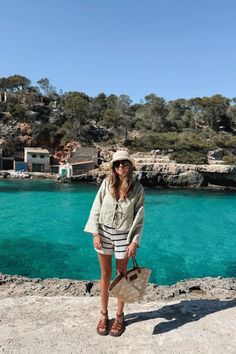 a woman standing on top of a cliff next to the ocean with a hat and bag