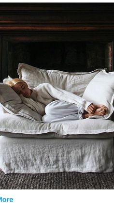 a woman laying on top of a bed covered in white sheets and pillows next to a fire place