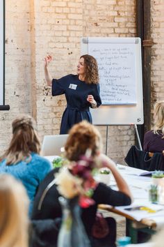 a woman standing in front of a whiteboard giving a presentation to people sitting at tables