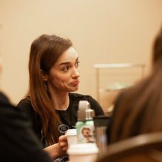 a woman is sitting at a table talking to another person who is holding a cup