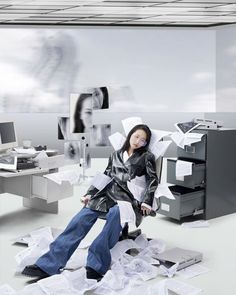 a woman sitting on top of a pile of paper next to a computer desk filled with papers