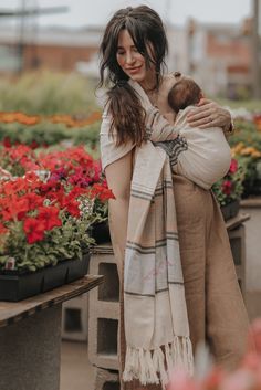 a woman holding a baby wrapped in a blanket while standing next to flowers and potted plants