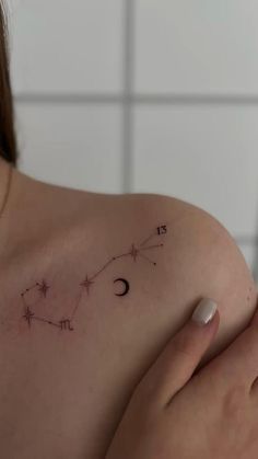 a woman's shoulder with the zodiac sign and stars on her left arm, in front of a white tile wall