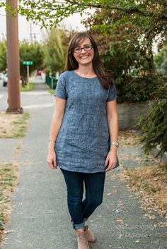 a woman standing on a sidewalk in front of a tree and wearing glasses with her hands in her pockets