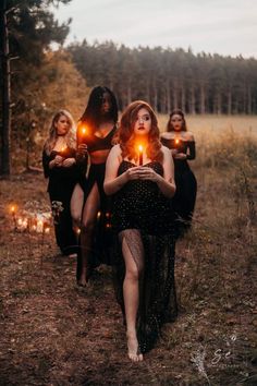 three women in black dresses are holding candles and walking through the woods with their faces lit up