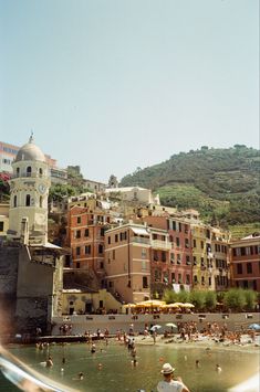 people are swimming in the water near some buildings and hills on top of a hill