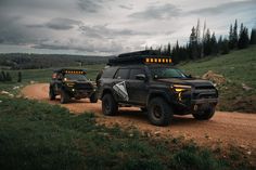 two vehicles driving down a dirt road in the mountains with trees and grass on both sides