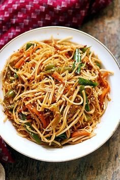 a white plate topped with noodles and vegetables on top of a wooden table next to a red napkin