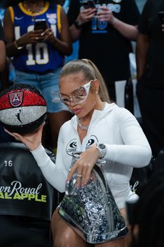 a woman in white shirt and glasses holding a basketball hat with other people behind her