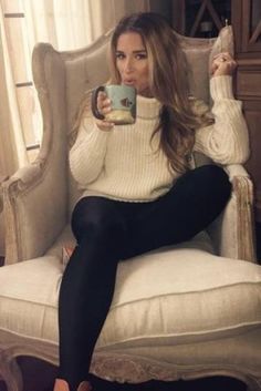 a woman sitting in a chair holding a cup and looking at the camera with her legs crossed