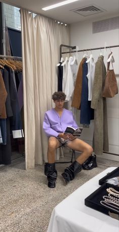 a man sitting in a chair reading a book next to a rack of clothes and shoes