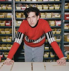 a man in a red sweater leaning over a table with boxes on the shelves behind him