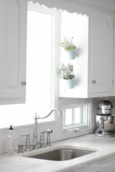 a kitchen with white cabinets and a sink in front of a window filled with potted plants