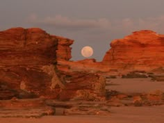 the moon is setting over some red rock formations in an arid area with sparse grass and rocks