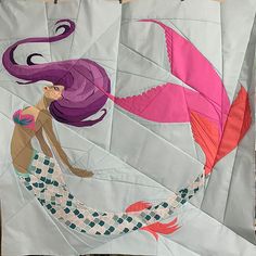 a piece of art that looks like a mermaid with long purple hair sitting on top of a quilt