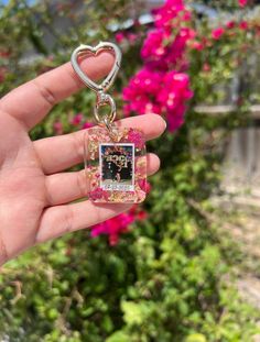 a hand holding a small square shaped keychain in front of some pink flowers