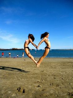 two women in bikinis jumping into the air on a beach with blue sky and water behind them