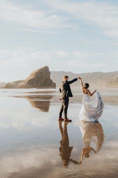 a bride and groom dancing on the beach with their reflection in the wet sand at low tide