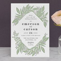 a white and green wedding card next to a vase with a flower in it on a table