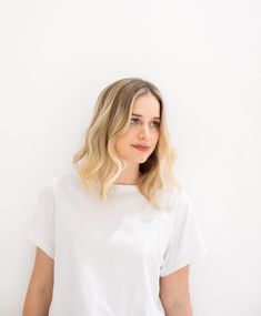 a woman standing against a white wall with her hands in her pockets and looking at the camera