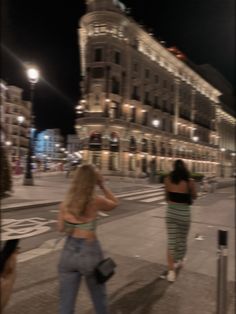 two women walking down the street in front of a large building at night with lights on