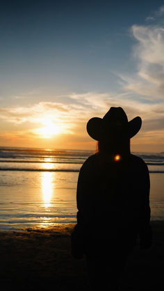 the silhouette of a person wearing a cowboy hat at sunset on the beach with water in the background