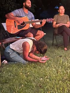 two men and a woman are sitting in the grass playing guitar