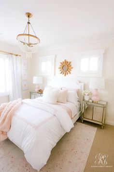 a white bed sitting in a bedroom next to a chandelier and two windows