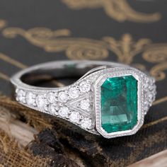 an emerald and diamond ring sitting on top of a piece of wood next to a book