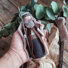 a hand holding an old fashioned doll in front of some green leaves and branches on a wooden table