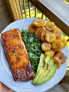 a white plate topped with meat and vegetables next to sliced avocado on a wooden deck