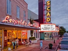 people are walking in front of a theater on the street at dusk with cars parked nearby