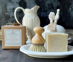 soap and other items are sitting on a plate next to a jug, brush, soap dish and box