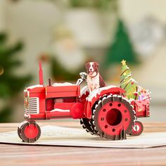 a dog riding on the back of a red tractor with a christmas tree in it