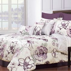 a bed with purple flowers on it in front of a white wall and large windows