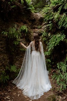 a woman in a white wedding dress standing on a path surrounded by trees and ferns