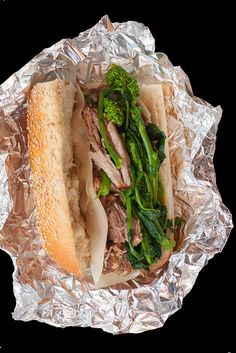 a sandwich with meat, broccoli and cheese wrapped in tin foil on a black background