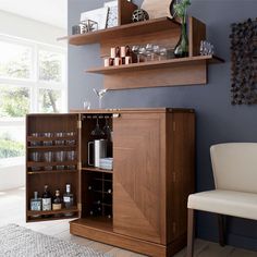 a wooden cabinet filled with lots of bottles and glasses next to a white chair in front of a window