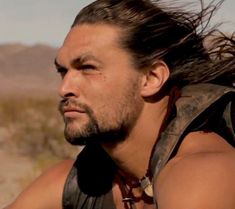 a man with long hair wearing a leather vest and looking off to the side in front of mountains