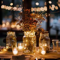 mason jars filled with flowers and lit candles are on a table in front of string lights