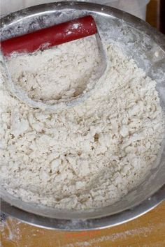 a metal bowl filled with white flour on top of a wooden table next to a red spatula