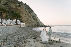 a bride and groom walking on the beach near some chaise lounges at sunset