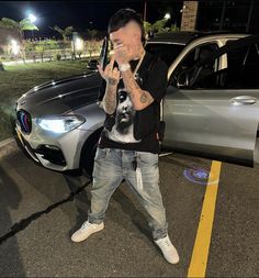 a man standing in front of a car making the peace sign with his hand while wearing jeans and sneakers