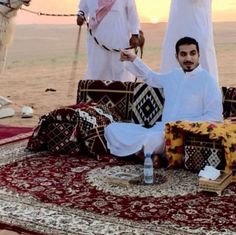 a man sitting on top of a rug next to two other men in white outfits