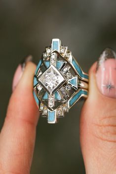 A close-up image of a woman's fingers holding a "Calliandra" design three-piece wedding ring set from Staghead Designs. This ring set features geometric Western-style design elements & a 2ct princess cut moissanite engagement ring with crushed turquoise inlays & diamond accents & is paired with two matching turquoise & diamond bands. This ring is shown in 14K white gold but can also be made in yellow or rose gold. Western Wedding Ring Stacker, Engagement Rings With Unique Stones, Unique Turquoise Rings, Navajo Wedding Rings, Southwestern Engagement Ring, Native Wedding Rings, Turqouise Wedding Ring, Southwest Engagement Ring, Tourquise Wedding Rings